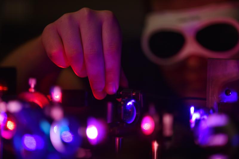 Eine Person trägt eine Schutzbrille und arbeitet in einem dunklen Labor an einem optischen Aufbau mit violetten und roten Laserstrahlen. Die Hand justiert präzise ein Linsensystem.