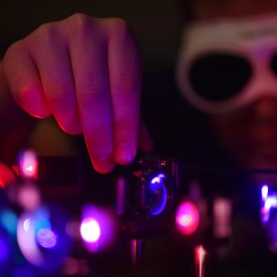 Eine Person trägt eine Schutzbrille und arbeitet in einem dunklen Labor an einem optischen Aufbau mit violetten und roten Laserstrahlen. Die Hand justiert präzise ein Linsensystem.